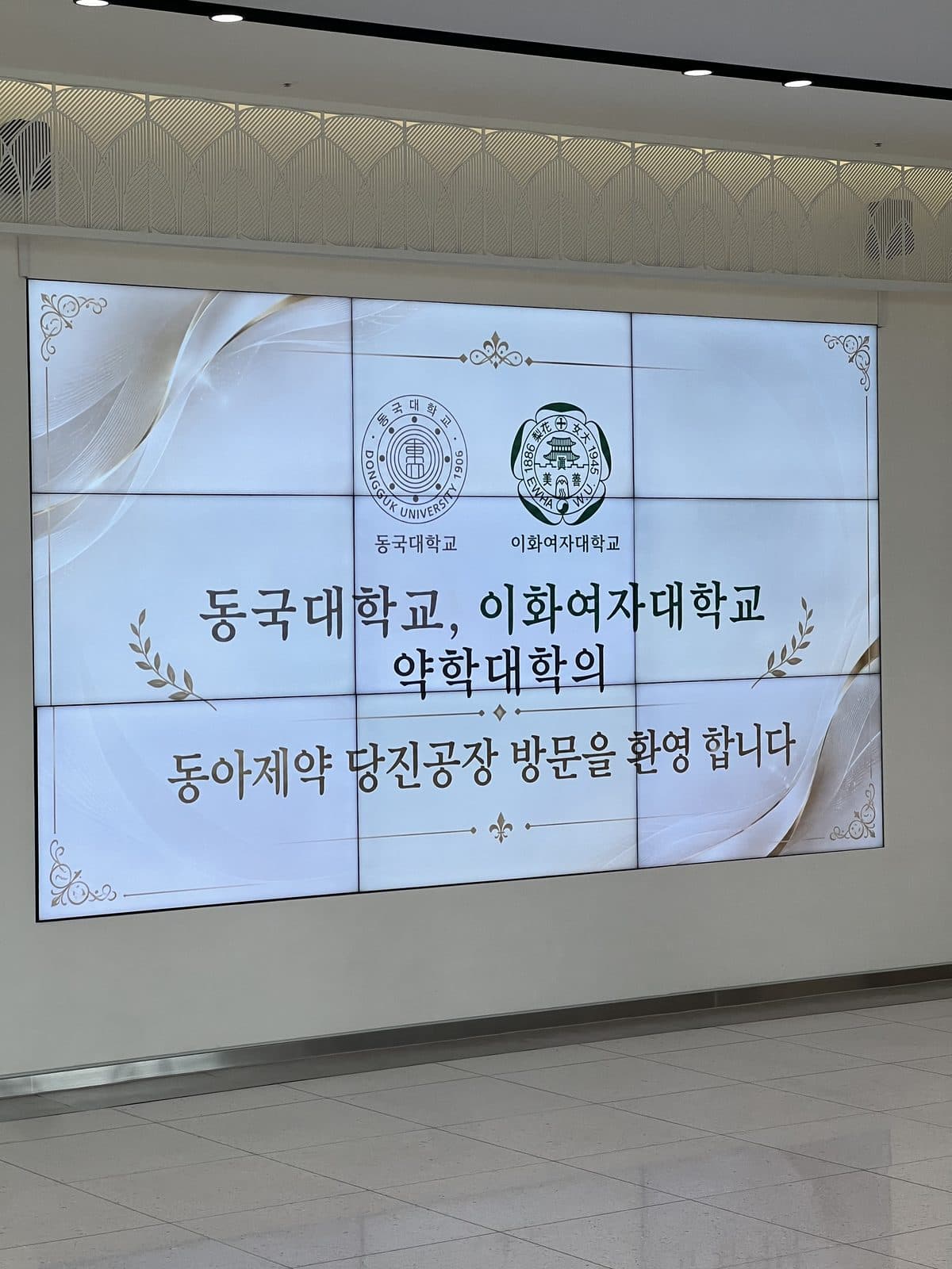 제약회사 공장 로비의 대형 화면에 동국대학교와 이화여자대학교 약학대학의 공장 방문을 환영하는 문구가 띄워져 있다.